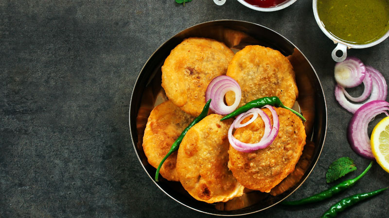 Freshly fried golden pyaaz kachoris topped with sliced red onions and green chilies served in a metal bowl