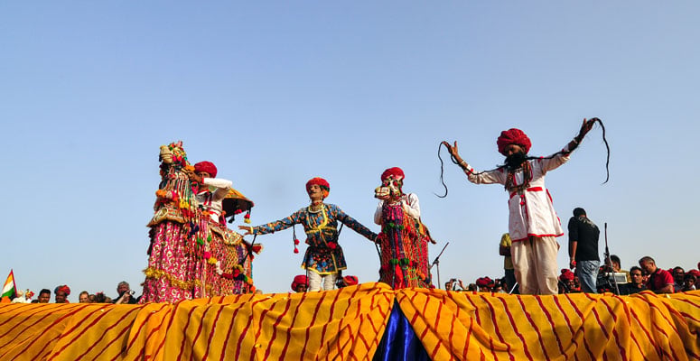 Folk artists in colorful traditional Rajasthani attire performing on stage against a blue sky